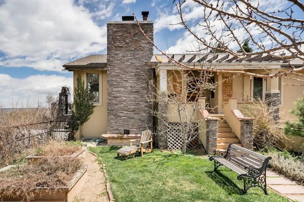 A house with a stone chimney, a covered porch, and a small garden with a bench and chair. - Expansive Luxury in Central Home With Downtown Views (Boulder)
