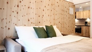 A bedroom with a wooden headboard, a bed with white and green pillows, a grey sofa, and a kitchen area visible through an open doorway.