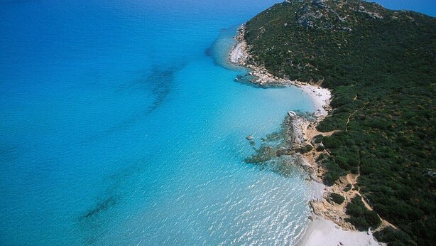 Vista aérea de una zona costera con playa, aguas azules cristalinas y una colina con una torre.