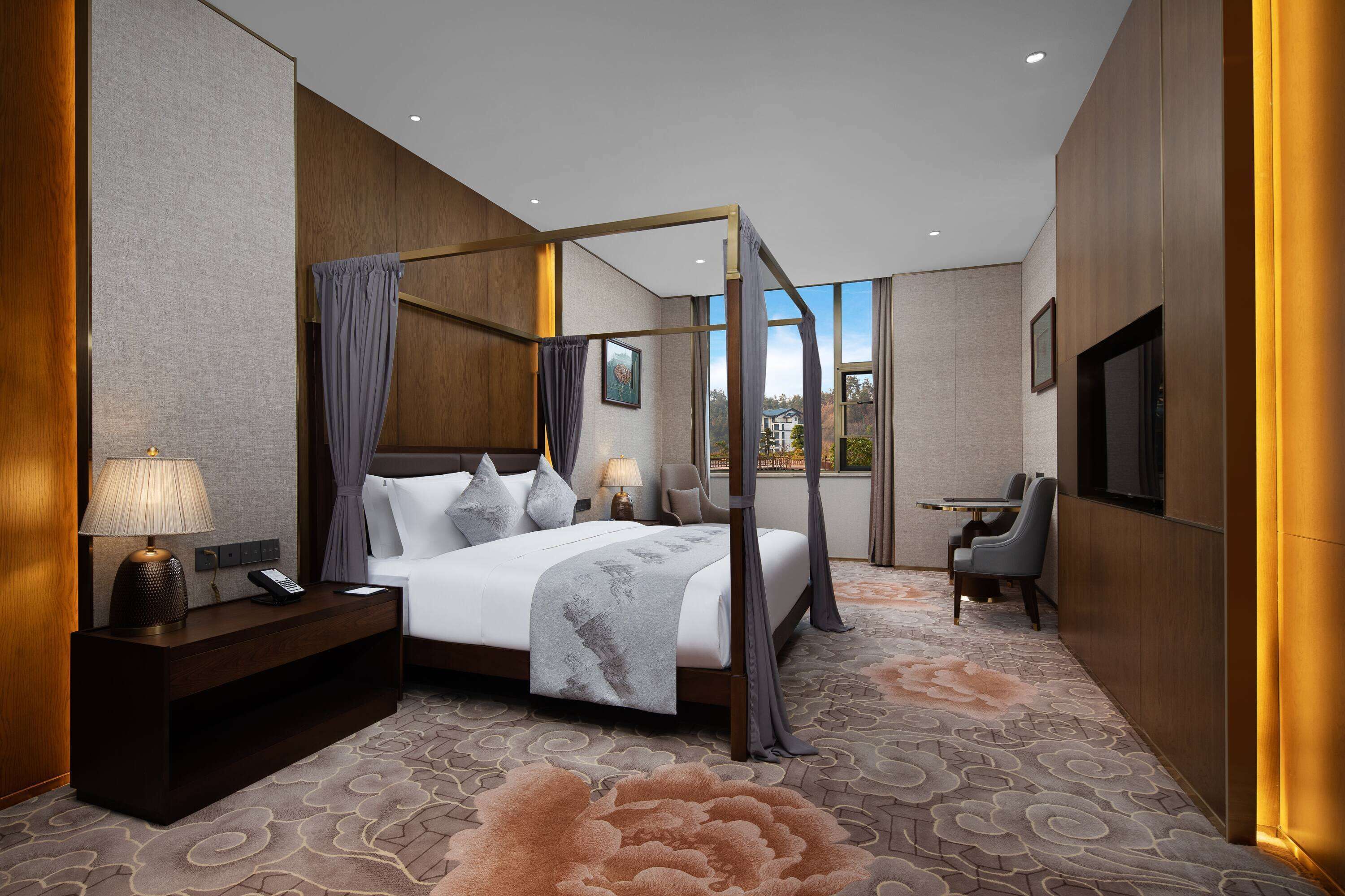 Une chambre d’hôtel moderne dotée d’un lit à baldaquin, d’une table de chevet avec une lampe, d’un bureau avec une chaise et offrant une vue sur l’extérieur grâce à de grandes fenêtres.