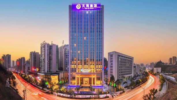Un hotel moderno de gran altura con una entrada destacada, rodeado de otros edificios y una calle transitada con estelas de luz.