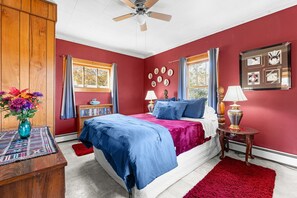 A bedroom with a bed, a nightstand, a lamp, a vase with flowers, and a window with curtains. - Whimsy Cottage - Perfect ski cottage & discounted lift tickets (North Troy)