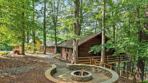 A rustic cabin surrounded by a forest, with a circular stone fire pit in the center. - Plenty of Water on the main channel. Perched on a bluff with breathtaking sunrise views over the lake, this remodeled 2BR/1BA log cabin offers memory-foam beds, well-stocked granite kitchen (farm-fresh eggs!), covered deck, stone fire pit, huge dock. (Union Hall)