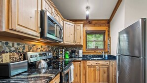 A compact kitchen with wooden cabinets, a stainless steel refrigerator, a microwave, a kettle, and a stone backsplash. - Plenty of Water on the main channel. Perched on a bluff with breathtaking sunrise views over the lake, this remodeled 2BR/1BA log cabin offers memory-foam beds, well-stocked granite kitchen (farm-fresh eggs!), covered deck, stone fire pit, huge dock. (Union Hall)