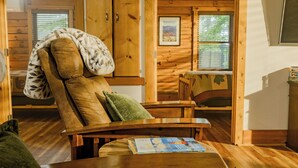 A wooden cabin interior with a recliner chair, a table with a magazine, and a window with blinds. - Plenty of Water on the main channel. Perched on a bluff with breathtaking sunrise views over the lake, this remodeled 2BR/1BA log cabin offers memory-foam beds, well-stocked granite kitchen (farm-fresh eggs!), covered deck, stone fire pit, huge dock. (Union Hall)