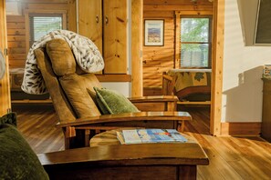 Interior d'una cabana de fusta amb una butaca relaxant, una taula amb una revista i una finestra amb persianes.