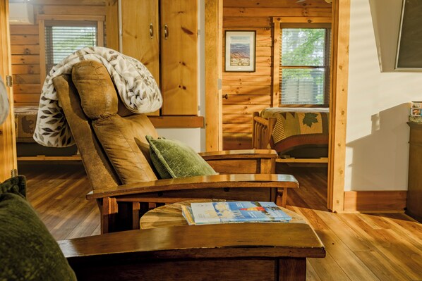 A wooden cabin interior with a recliner chair, a table with a magazine, and a window with blinds. - Plenty of Water on the main channel. Perched on a bluff with breathtaking sunrise views over the lake, this remodeled 2BR/1BA log cabin offers memory-foam beds, well-stocked granite kitchen (farm-fresh eggs!), covered deck, stone fire pit, huge dock. (Union Hall)