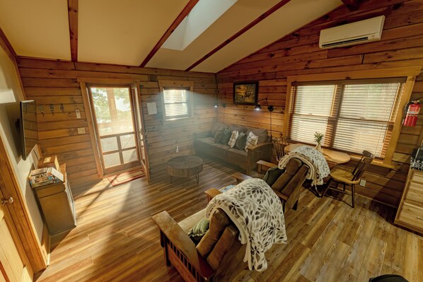 A wooden cabin interior with a sofa, chairs, a small table, and a kitchen area. - Plenty of Water on the main channel. Perched on a bluff with breathtaking sunrise views over the lake, this remodeled 2BR/1BA log cabin offers memory-foam beds, well-stocked granite kitchen (farm-fresh eggs!), covered deck, stone fire pit, huge dock. (Union Hall)