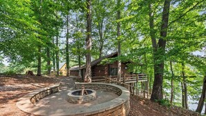 A rustic cabin surrounded by trees, a circular stone fire pit, and a wooden deck. - Main Channel-Lots of Water-PickleBall-Hot Tubs-Fire Pits-2 Private Docks. Exclusive use of all 5 log homes, 8 acres, 1300 ft shoreline. Sleeps 36. 1400 sq ft venue, events to 75 (fee applies): weddings, reunions, retreats, milestones. 4.9★ Superhost (Union Hall)