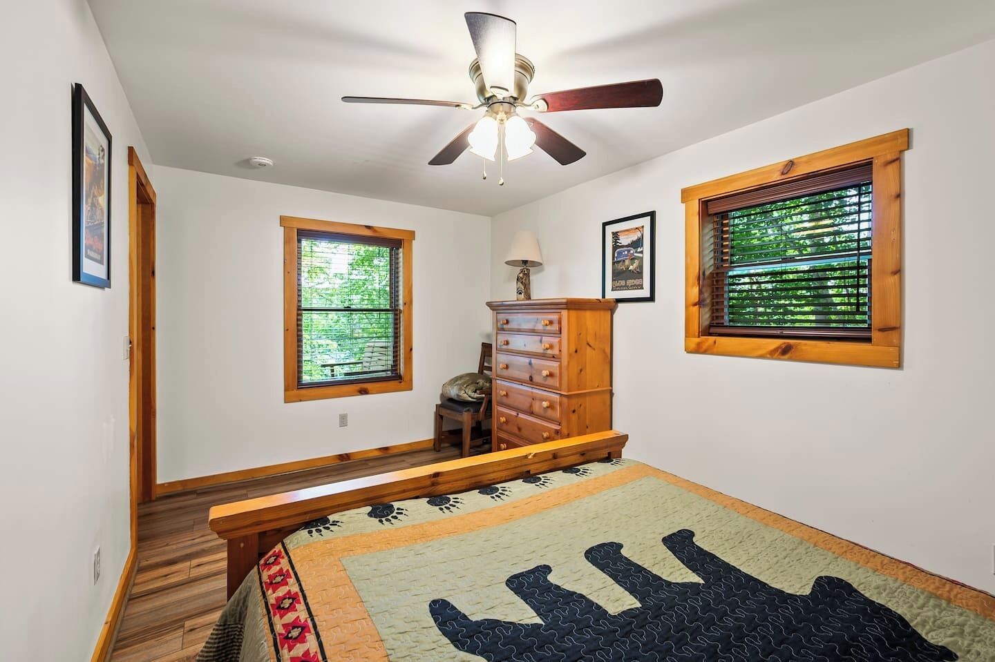 Een slaapkamer met een bed, een houten dressoir, twee ramen met zonwering, een plafondventilator en een trap.