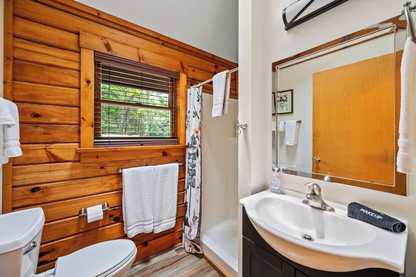 Une salle de bain avec un mur en bois, une fenêtre avec des stores, un rideau de douche, un lavabo avec un miroir et des toilettes.