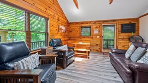 A log cabin interior with a wooden floor, leather sofas, a wooden dining table, and large windows with blinds.