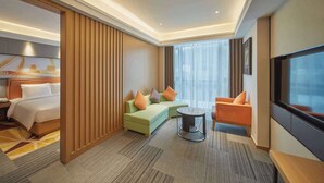Une chambre d’hôtel moderne avec un lit, un canapé vert agrémenté de coussins orange, une table basse ronde et un téléviseur.