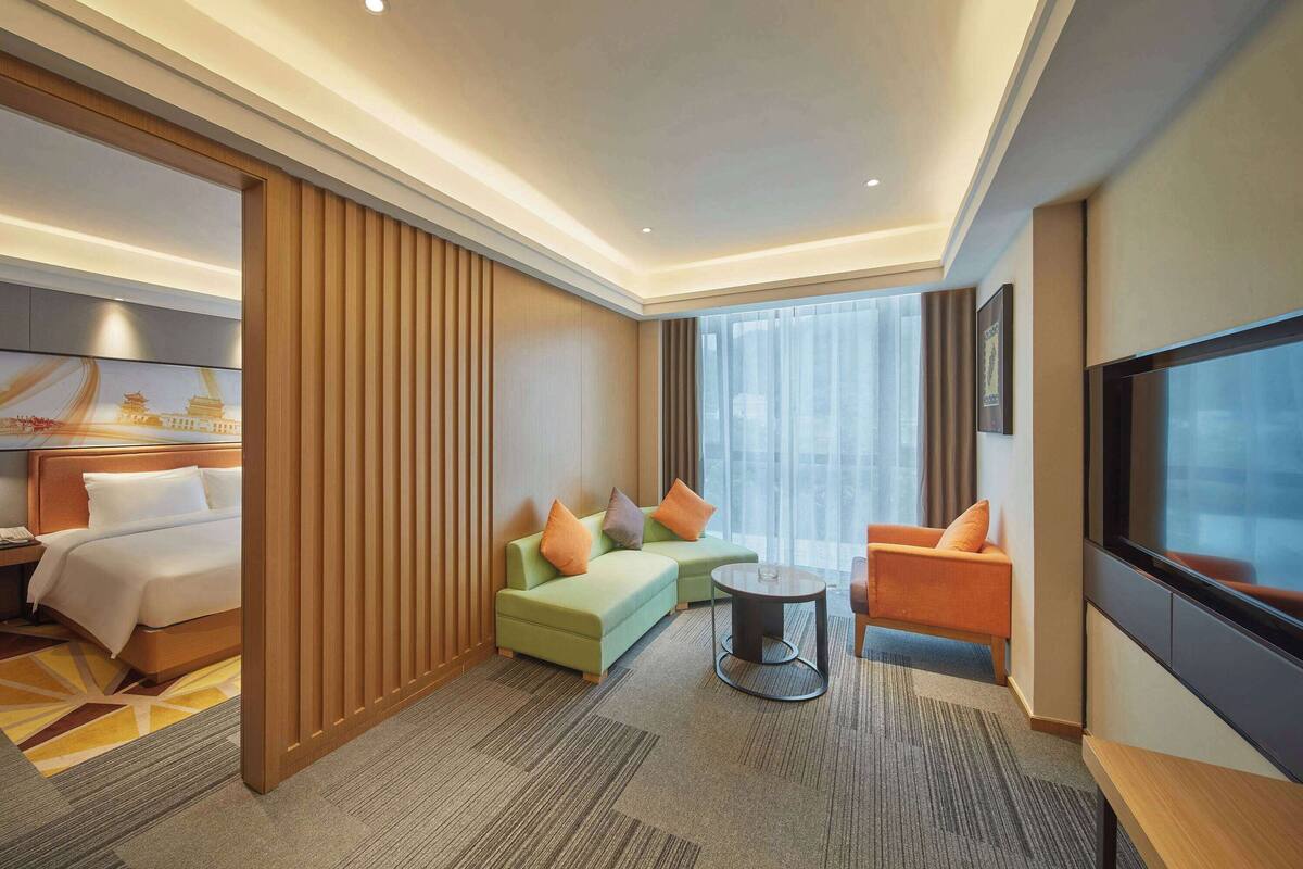 A modern hotel room with a bed, a green sofa with orange cushions, a round coffee table, and a TV.