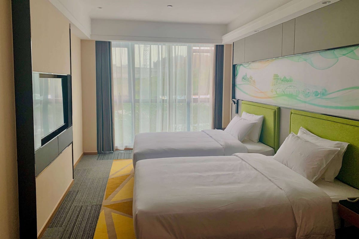 A hotel room with two beds, a flat-screen TV, a large window with curtains, and a painting on the wall.