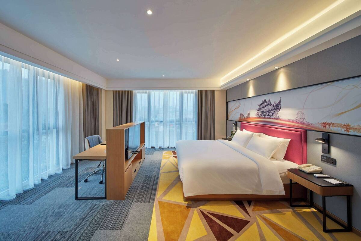 A hotel room with a bed, a desk, a chair, a TV, and a large window with curtains.