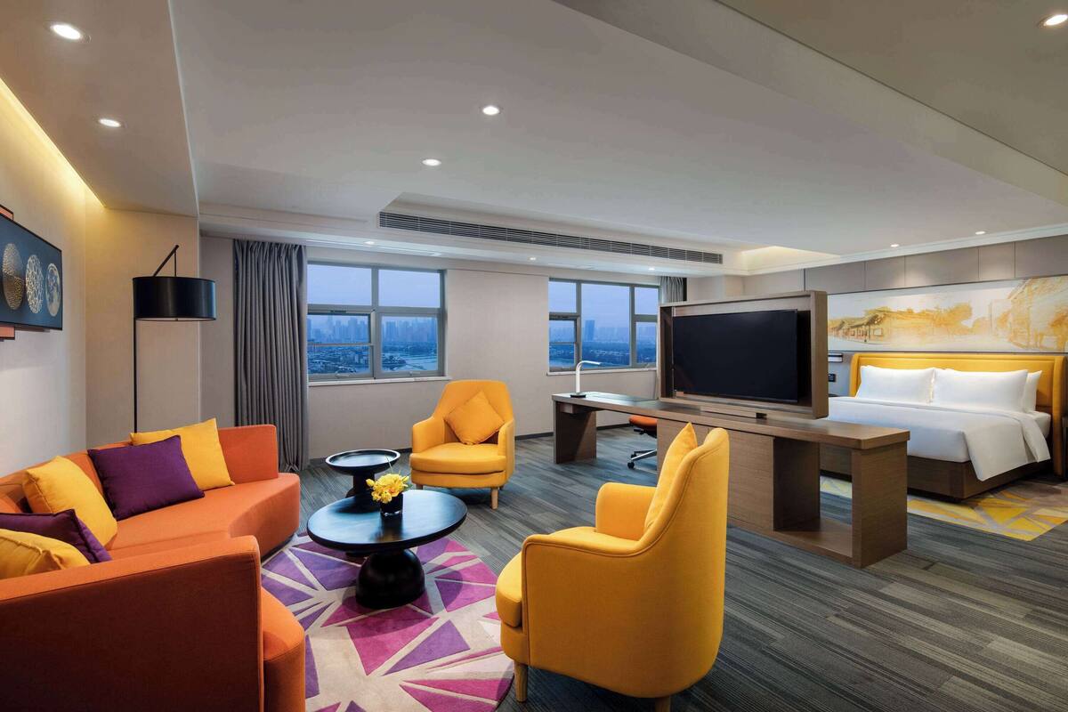 A modern hotel room with a sofa, armchairs, a desk, a bed, a TV, and large windows offering a city view.