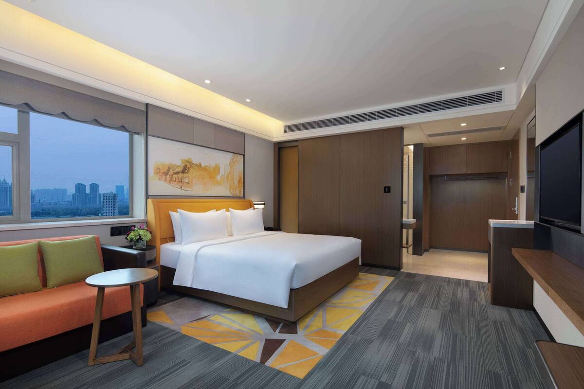 A hotel room with a large bed, a sofa, a small table, a TV, and a city view.