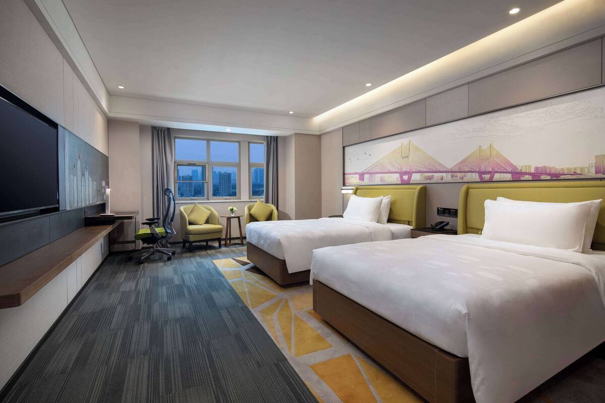 A hotel room with two beds, a desk, a chair, a TV, and a large window with a cityscape view.