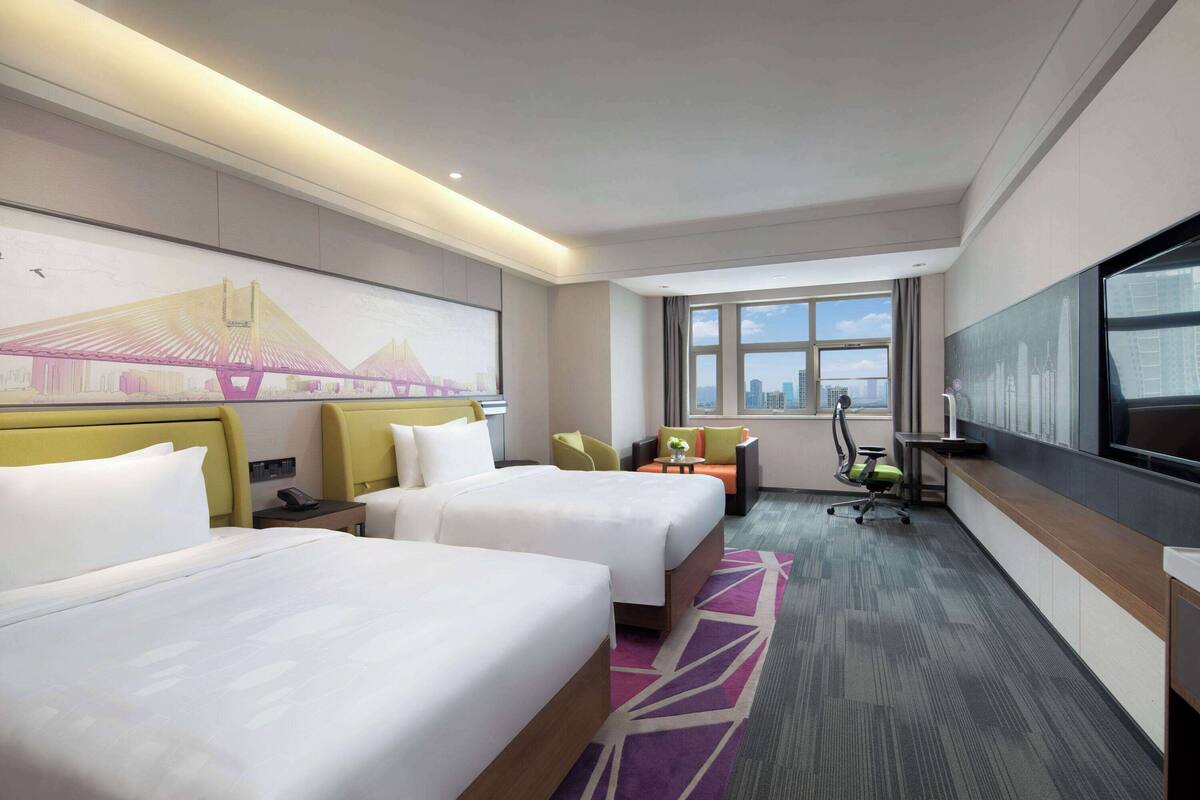 A hotel room with two beds, a desk, a chair, a TV, and a large window with a city view.