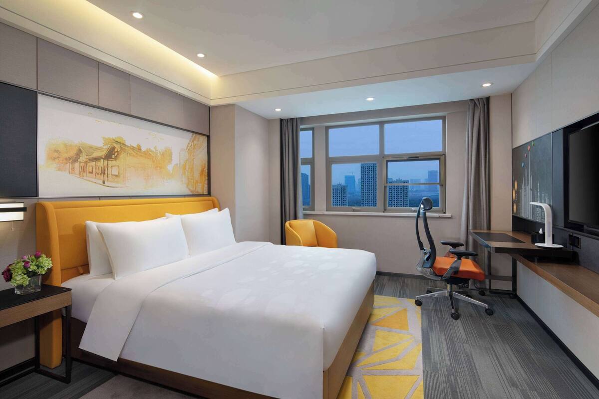 A hotel room with a large bed, a desk with a chair, a television, and a window with a city view.