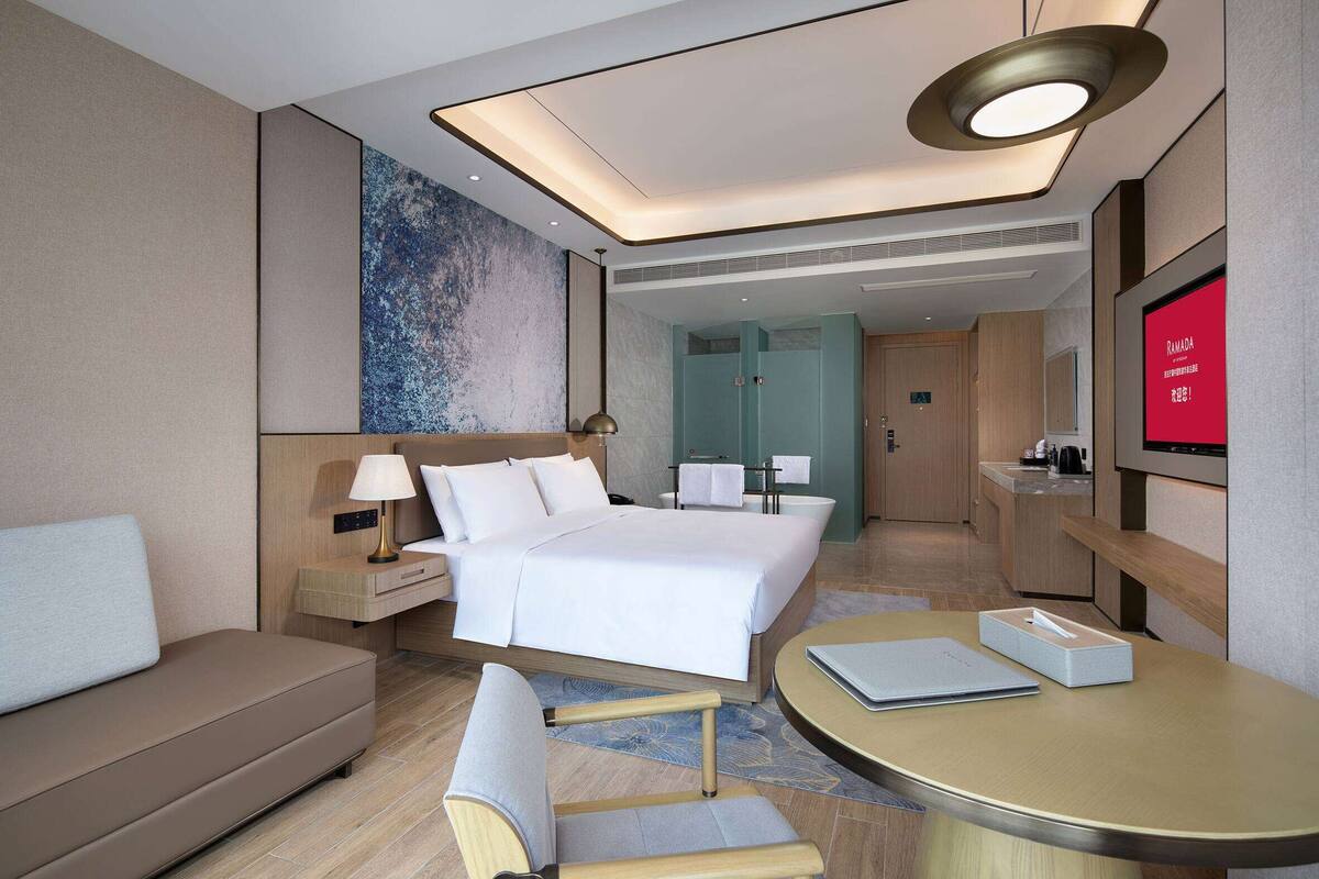 Een moderne hotelkamer met een bed, nachtkastje, lamp en een televisie waarop 'Ramadan' staat.