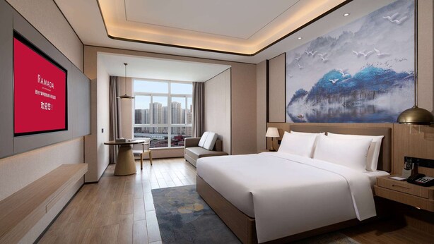 Ein modernes Hotelzimmer mit einem großen Bett, einem Schreibtisch, einer Couch und Blick auf die Stadt durch das Fenster.