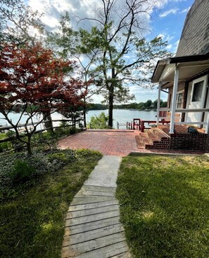 A wooden deck leading to a brick patio with a view of a lake and a house with a porch. - Waterfront With Relaxing Views (Pasadena)