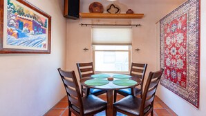A dining area with a round table, four chairs, a painting on the wall, a window with blinds, and a patterned rug.