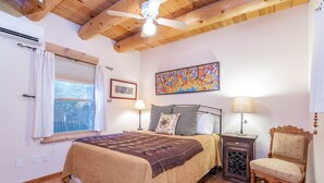 A bedroom with a bed, a chair, a nightstand, a ceiling fan, a window with curtains, and a painting on the wall.