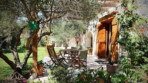 An outdoor seating area with a table and chairs, surrounded by greenery and a stone wall. - Traditional Cretan stone cottage on the semi-rural outskirts of Kefalas, Vamos (Kefalas)