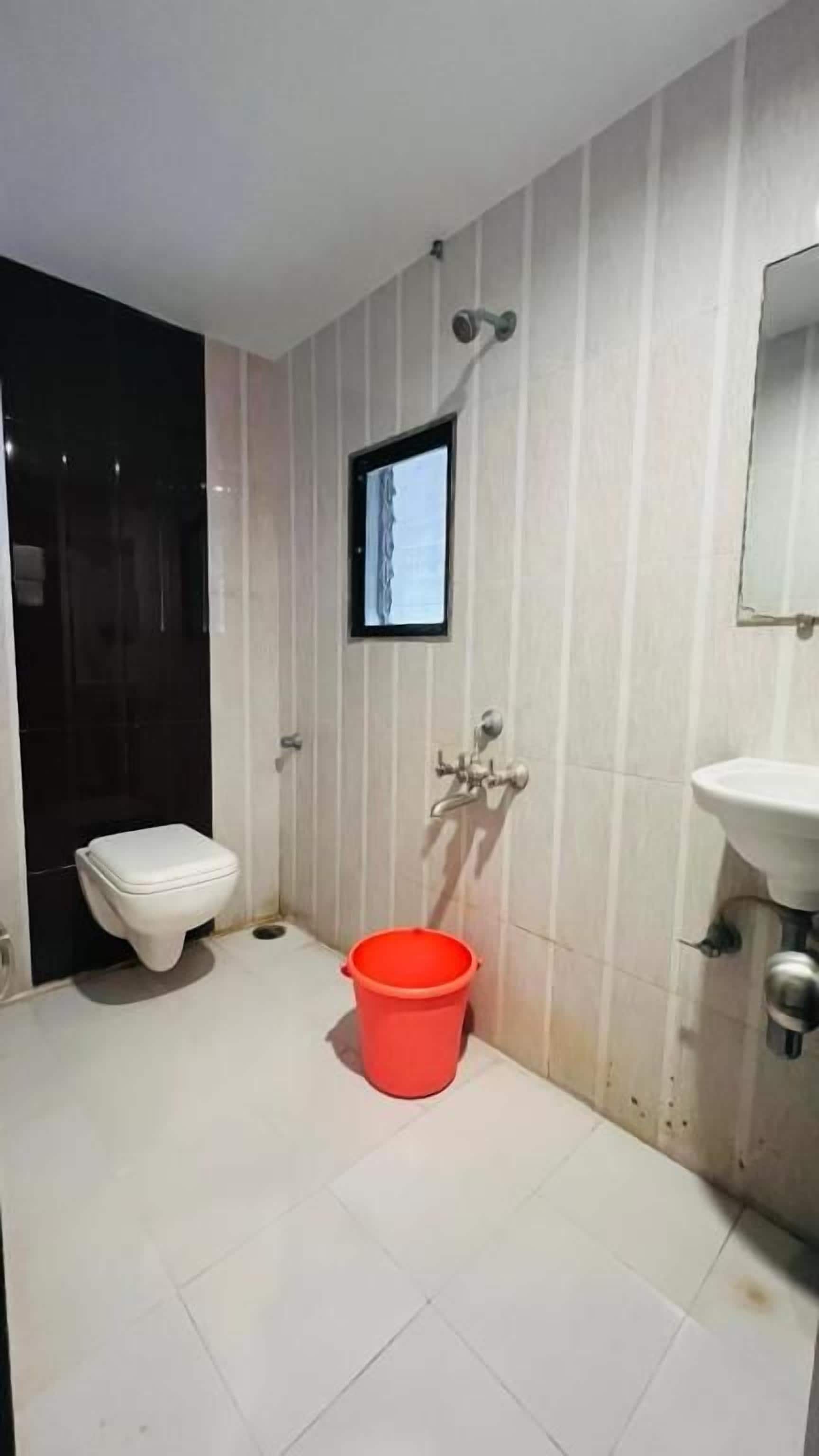 Un baño con inodoro, lavamanos, un cubo rojo y una pantalla pequeña montada en la pared.