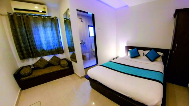 A bedroom with a bed, a small seating area, an air conditioning unit, and a door leading to a bathroom. - Farmhouse Residency (Vasai)