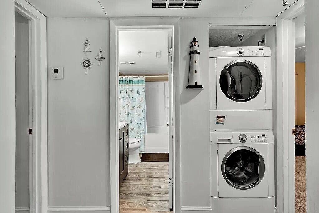 A laundry room with two stacked front-loading washing machines, a vent on the ceiling, and a door leading to a bathroom with a toilet and a shower curtain.