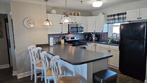 A kitchen with white cabinets, a black refrigerator, a microwave, and a stove. There are two pendant lights, a clock, and a window with a plaid curtain. - Incredible Oceanfront End-Unit Condo with Pool & Elevator Access (North Topsail Beach)