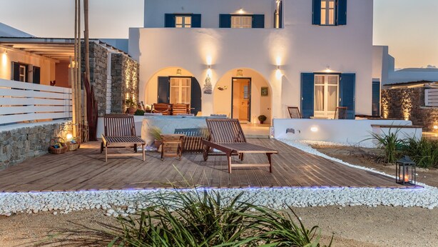 A modern white building with blue shutters, a wooden deck with lounge chairs, and a stone wall.