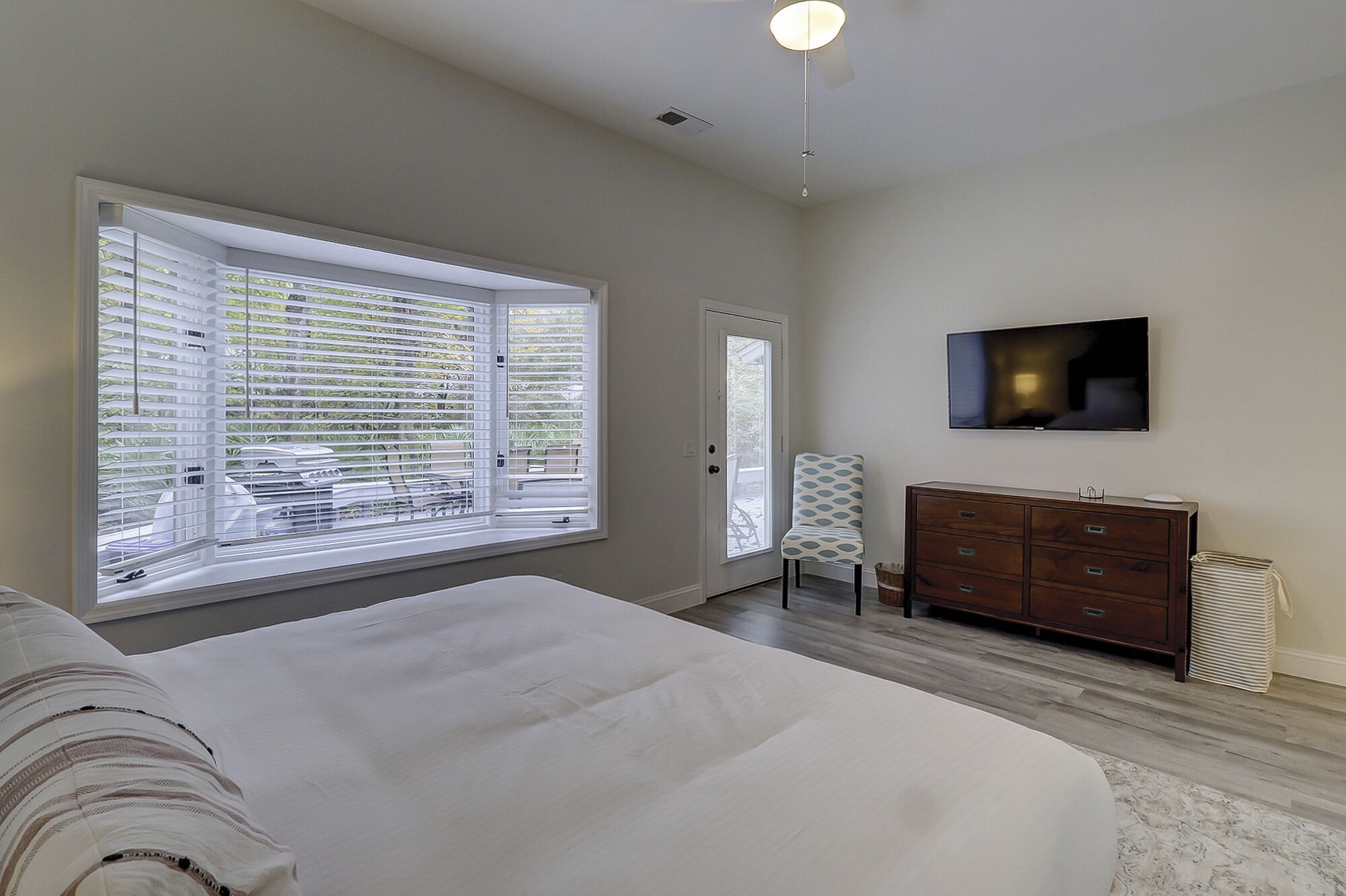 A bedroom with a large window, a bed, a dresser, a chair, and a TV mounted on the wall.