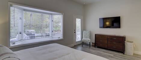 A bedroom with a large window, a bed, a dresser, a chair, and a TV mounted on the wall.