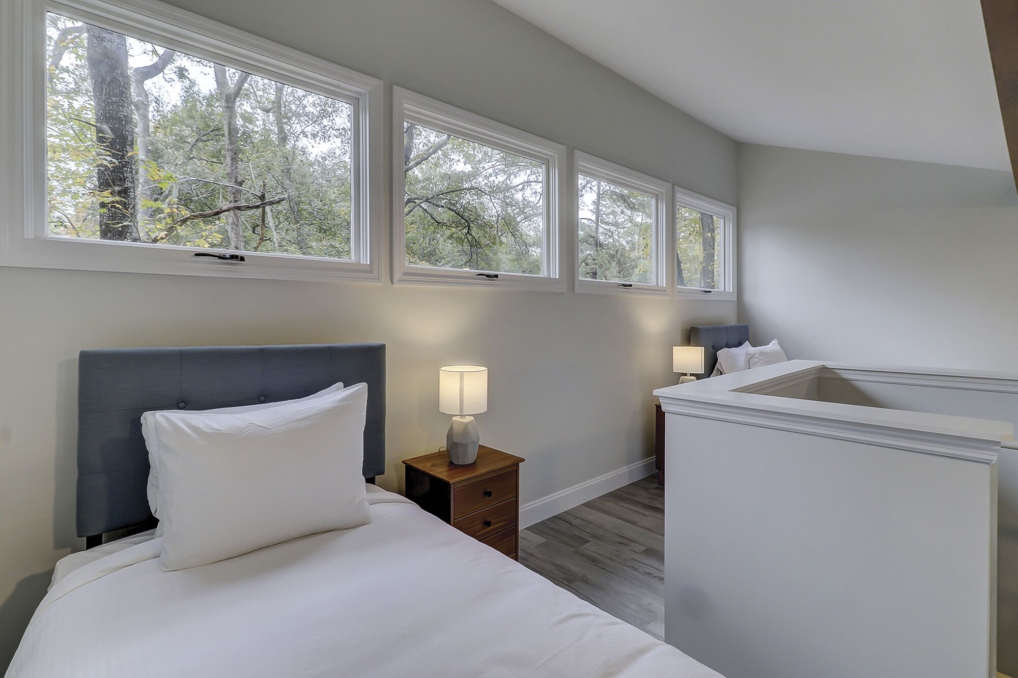 A bedroom with a bed, bedside tables, and a nightstand. There are three windows providing views of trees.