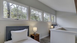Un dormitorio con cama, mesitas de noche y mesilla auxiliar. Hay tres ventanas que ofrecen vistas a árboles.