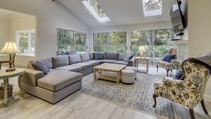 Amplia sala de estar con un sofá modular, una mesa de centro y un tapete estampado. Grandes ventanales permiten la entrada de luz natural y vistas al verde exterior.