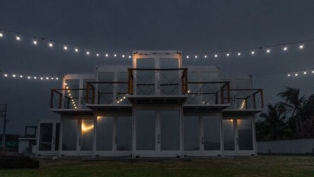 Un edificio moderno de varios niveles y amplios ventanales, rodeado de césped y luces de cadena.