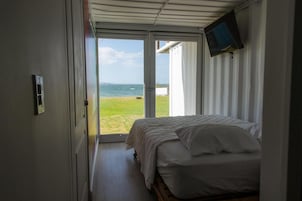Un dormitorio con cama, televisor montado en la pared y puerta que da a un balcón con vista al mar.