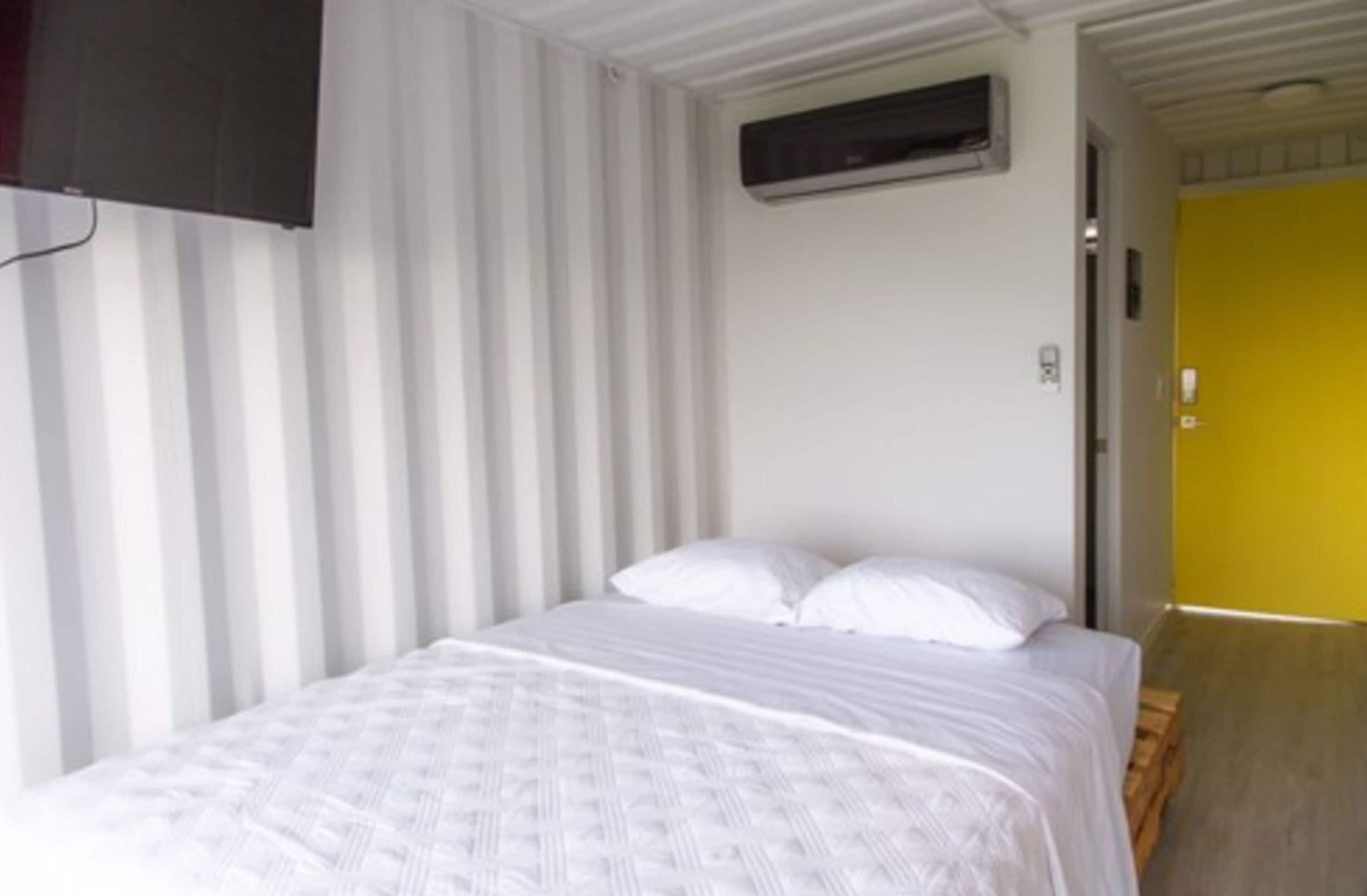 Una habitación con una cama, cortinas blancas, una puerta amarilla y un equipo de aire acondicionado instalado.