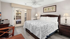Un dormitorio con cama, escritorio, silla, mesita de noche, lámpara, ventilador de techo y una puerta que da a un balcón.