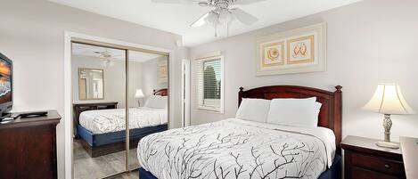 A bedroom with a bed, a nightstand, a lamp, a ceiling fan, a mirror, and artwork on the wall.