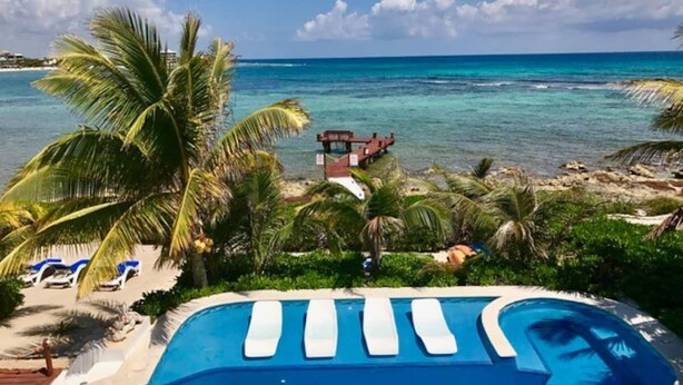 Piscina com espreguiçadeiras e palmeiras, com vista para a praia e o oceano.