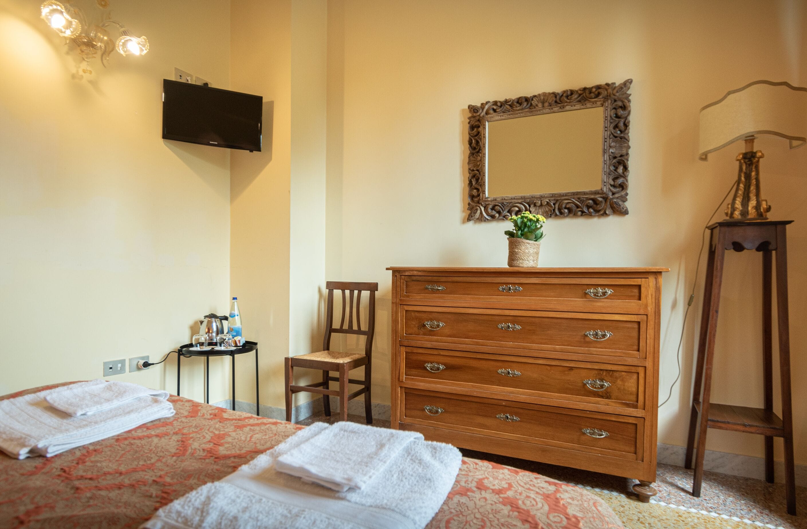 A hotel room with two beds, a wooden dresser, a mirror, a small table with a plant, and a lamp.