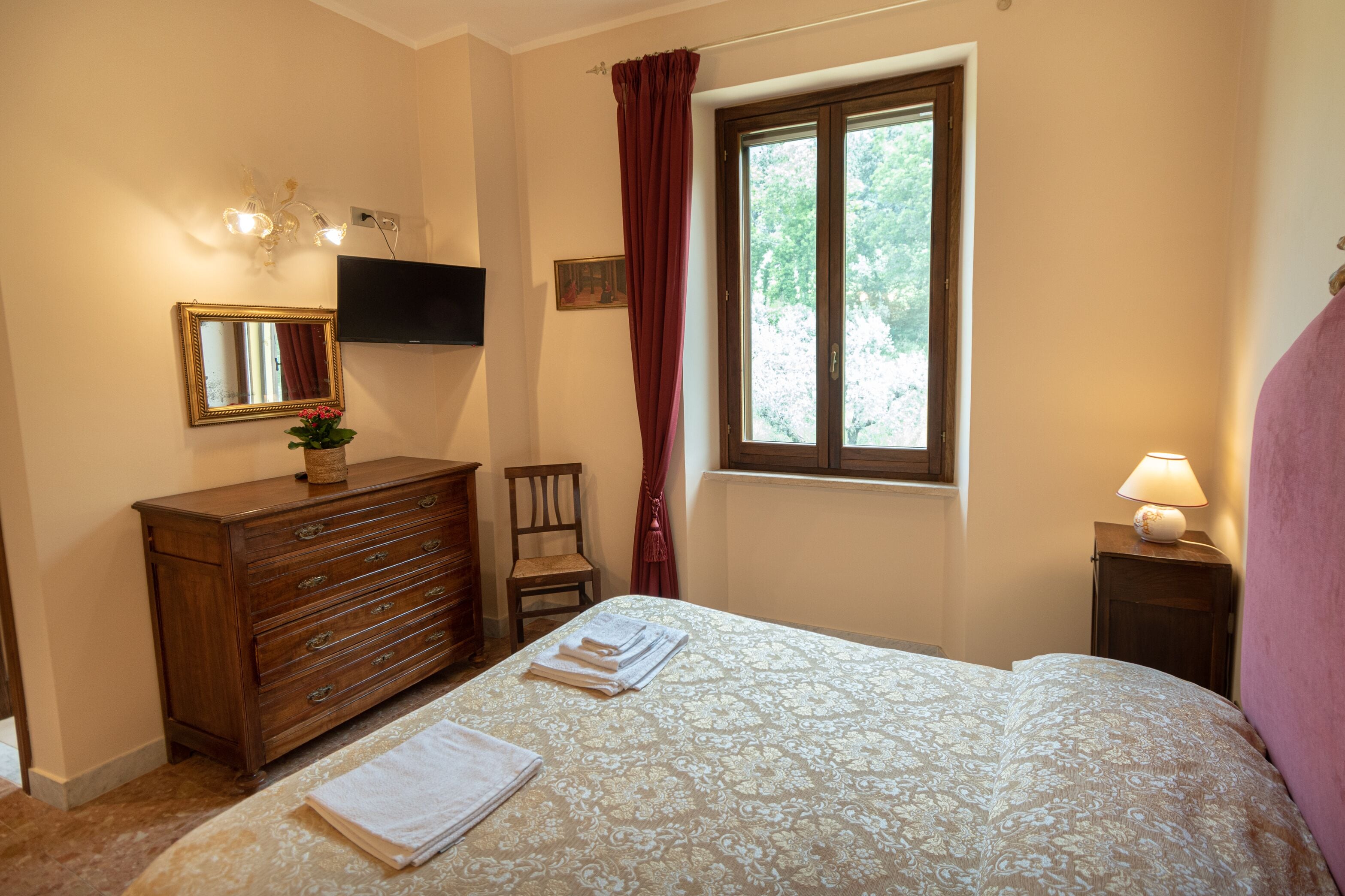 A bedroom with a bed, a dresser, a chair, a window with curtains, a lamp, and a TV mounted on the wall.