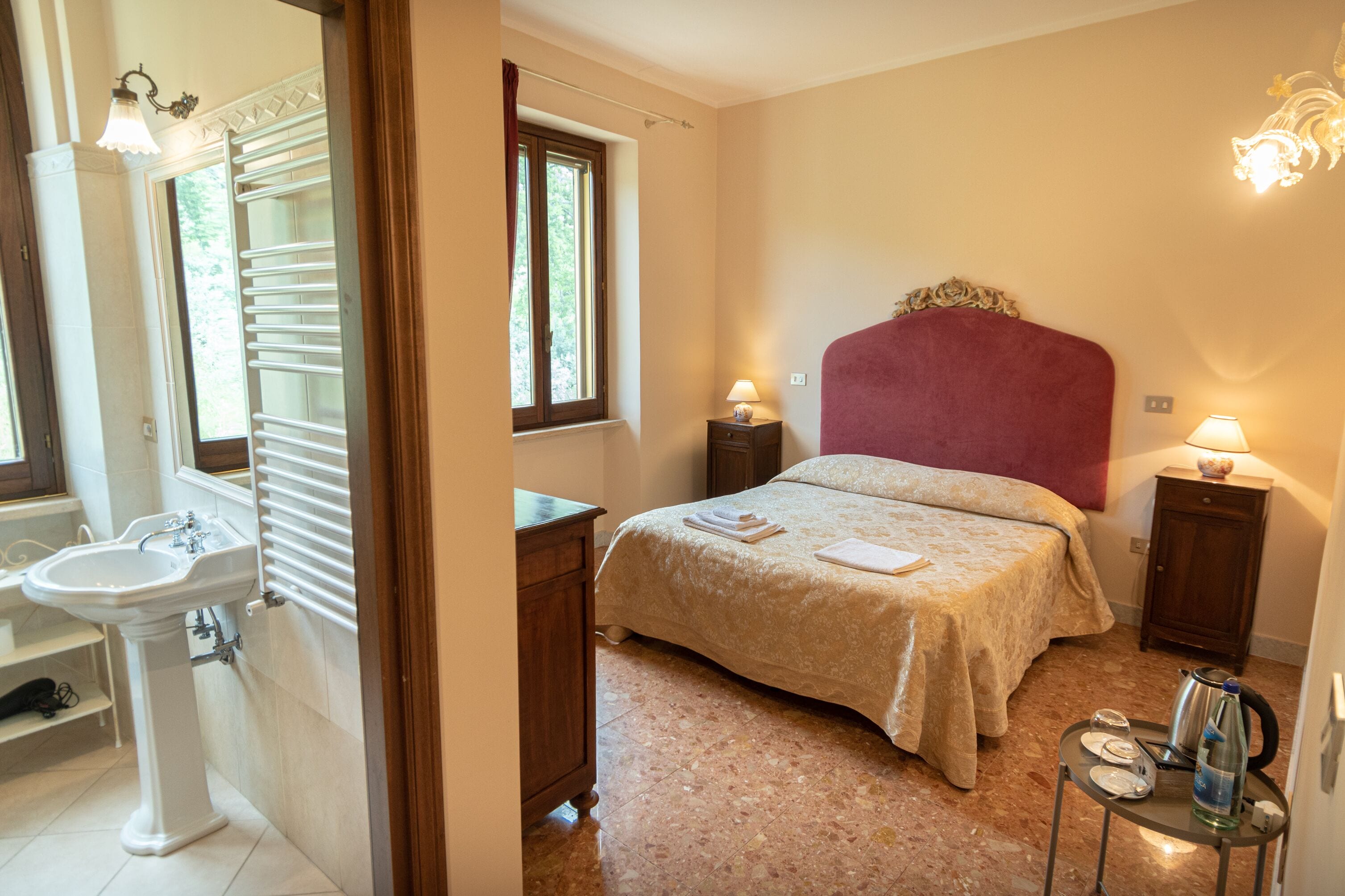 A bedroom with a bed, bedside tables, a nightstand, a window, a radiator, and a bathroom with a sink and mirror.